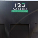 Déploiement multi-sites : logo 123 Pare-Brise imprimé sur panneau pour un bâtiment en bardage noir à Décines.